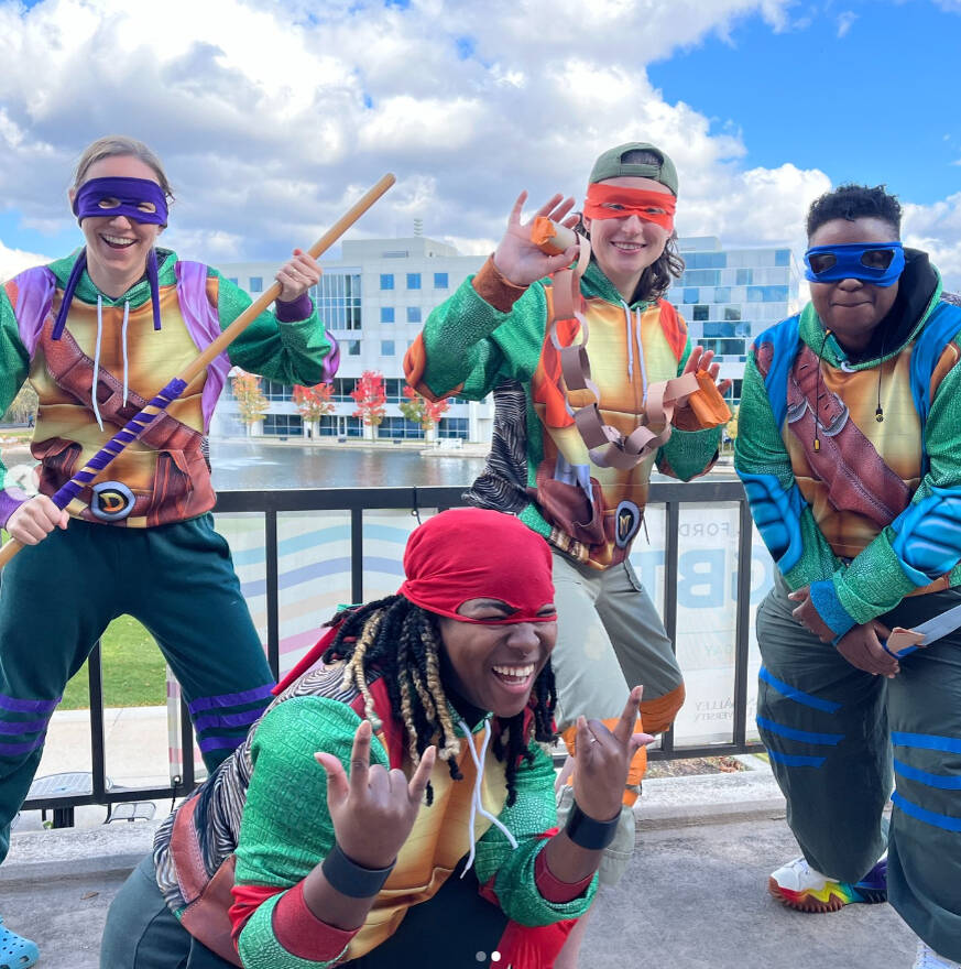 Four people dressed as the Teenage Mutant Ninja Turtles pose enthusiastically outdoors on the GVSU campus. Wearing colorful turtle-themed outfits and purple, orange, blue, and red masks, they smile, hold props, and make playful gestures toward the camera.
